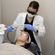 A female dermatologist wearing a lab coat and protective eyewear performs a facial laser treatment on a female patient, who is lying down with eye covers and a blanket.