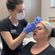 A female healthcare professional wearing gloves and a surgical mask is injecting a syringe into the forehead of a female patient, who is sitting back in a medical chair.