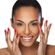 A person with a joyful expression has their hands near their face, showing off red-painted nails. They have dark hair pulled back and are smiling broadly. The background is white.
