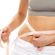 A person in a gray crop top and white bottoms measures their waist with a measuring tape against a white background. The person holds one end of the tape in each hand, wrapping it around their bare midriff.