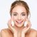 A young woman with a radiant smile, light makeup, and blonde hair pulled back, gently touching her face with her fingertips against a white background.