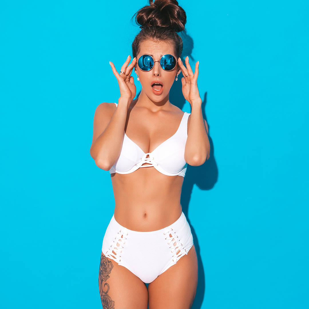 A woman in a white bikini stands in front of a bright blue background. She is wearing round blue sunglasses, with her mouth open in a surprised expression and her hands adjusting the sunglasses. Her hair is styled in a high bun.