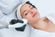 A woman in a white spa robe lies with her eyes closed while a gloved technician applies a laser treatment device to her face. The setting appears to be a professional skincare or medical spa facility.