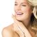 A person with long, wavy blonde hair, smiles brightly while looking slightly to the side. They have pink lips, rosy cheeks, and are resting their chin on their hands. The background is plain white, emphasizing their radiant complexion and happy expression.