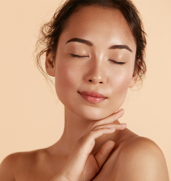 A woman with closed eyes gently touches her neck with one hand against a soft peach-colored background. She has a serene expression and her makeup is natural and minimal. Her hair is pulled back, and her skin looks smooth and glowing.