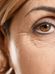 Close-up of the right eye of an older person with light brown hair. The image shows detailed wrinkles around the eye and eyebrow, highlighting signs of aging. The background is plain and bright.