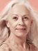 A close-up portrait of an elderly woman with a serene expression and long, gray hair, set against a soft peach background. She is wearing a subtle smile and a strap of a top is visible on her shoulder.