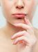 A close-up of a person slightly biting their bottom lip, with one hand gently touching their lips. The person has a light complexion and is wearing pale pink nail polish. The background is plain and neutral.