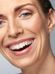 Close-up of a smiling woman with fair skin, showing her white teeth. Her dark hair is pulled back, and she has minimal makeup, highlighting her natural beauty against a light gray background.