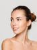 A woman with light skin and brown hair tied in a bun looks to her left, smiling slightly. She has bare shoulders and is set against a plain grey background. Her expression is calm and content.