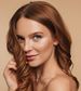 A woman with long, wavy, auburn hair poses against a beige background. She is looking at the camera with a slight smile, holding a strand of hair in her hand. Her complexion is fair and her makeup is minimal, featuring natural tones.