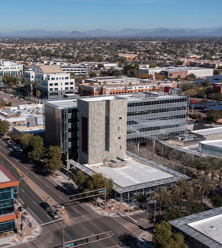 Aerial view of a modern, multi-story building with large glass windows and stone facade surrounded by an urban landscape. Streets with vehicles and palm trees run alongside the building. Mountains are visible in the distant background under a clear blue sky.