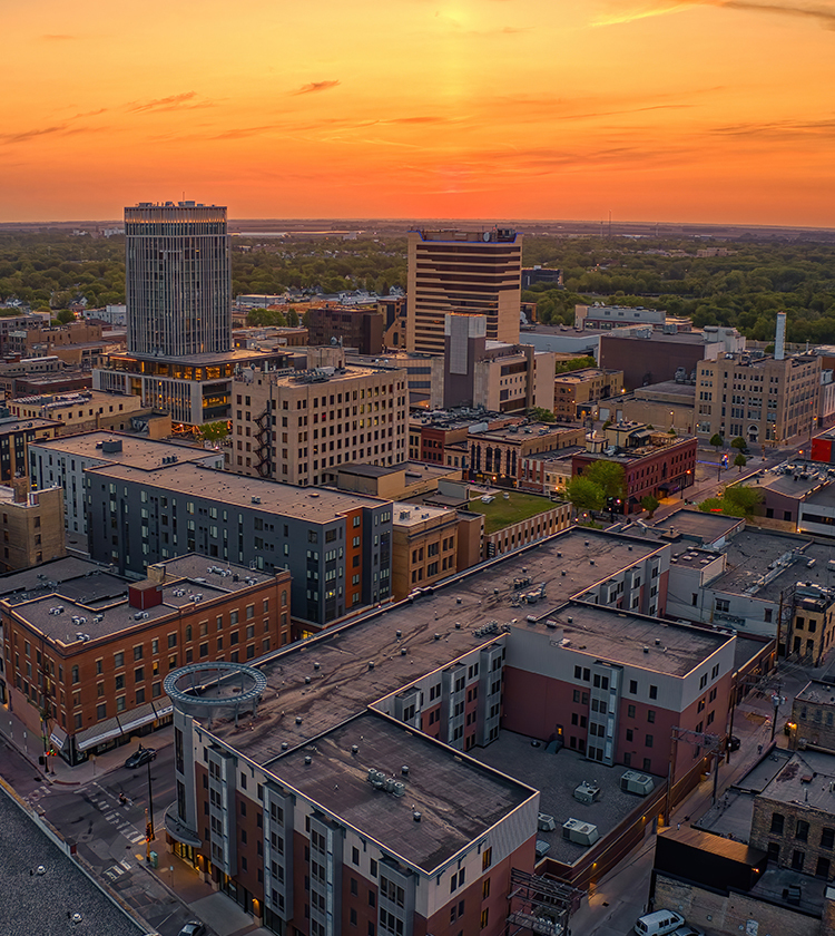 Aerial view of an urban landscape at sunset. The sky is painted in warm tones of orange and yellow. Numerous buildings, some tall and some low-rise, are visible with a mix of modern and historic architecture. Trees and greenery are interspersed throughout.