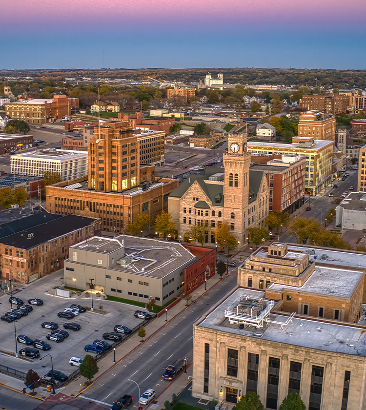 Aerial view of a mid-sized city at sunset, featuring a mix of historic and modern buildings. In the foreground, a clock tower stands prominently. Parking lots and tree-lined streets run between the buildings. The sky transitions from pink to blue on the horizon.