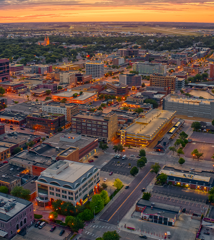 Aerial view of a cityscape at sunset, with lights illuminating streets and buildings. The horizon boasts a colorful sky with hues of orange, pink, and purple. Trees and parked cars are visible in various parking lots among the densely packed urban structures.