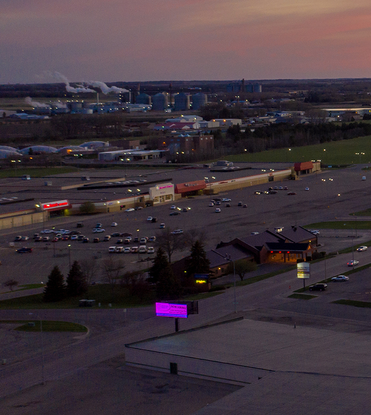 A view of a shopping center with several parking lots, some filled with vehicles. There are commercial buildings including a restaurant with a red roof. In the background, industrial structures emit smoke, and the sky is tinged with colors of the setting sun.