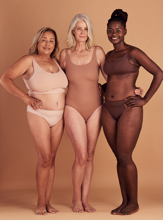 Three women of different ages and body types stand together in neutral-toned underwear against a beige background, smiling confidently. The woman on the left is wearing a two-piece, the woman in the center is in a one-piece, and the woman on the right is in a two-piece set.