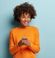 A cheerful young woman with curly hair, wearing an orange sweater, holding a smartphone and looking away with a happy expression, against a light blue background.