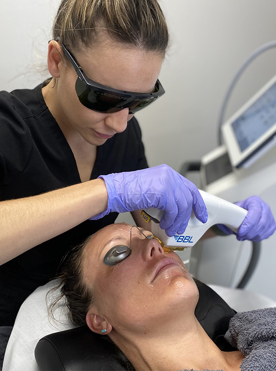 A woman is receiving a facial laser treatment from a technician wearing safety goggles and purple gloves. The patient has protective eyewear on and is lying on a clinic bed while the technician uses a handheld laser device on her face.