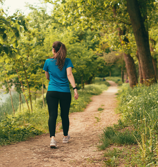 A woman in a blue top and black leggings is jogging on a dirt path surrounded by lush green trees.