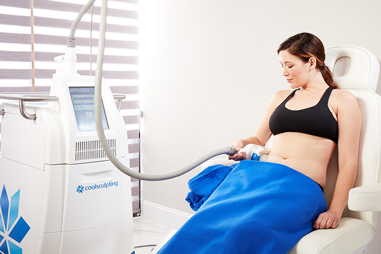A woman in a sports bra and blue covering sits in a medical chair, undergoing a non-invasive coolsculpting procedure on her abdomen, with a machine beside her displaying data.