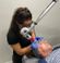 A female medical professional wearing glasses and blue gloves uses a laser treatment device on a patient's face in a clinical setting.
