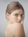 A young woman with fair skin and blue eyes looks over her bare shoulder. She has light brown hair tied back and wears no visible clothing or accessories. The background is a plain, light gray, creating a simple and minimalist portrait.