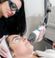 A woman is undergoing a skin treatment with laser equipment in a clinic. She is lying down with protective goggles on. The clinician, also wearing goggles and gloves, is holding the laser device close to the patient's face.