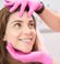 A person with brown hair smiles while receiving a skincare or medical treatment. Someone wearing bright pink gloves gently touches their forehead and chin. The setting appears to be a clinic or spa.