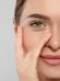 Close-up of a person touching their face with one hand. The person has green eyes, clear skin, and is slightly smiling with light makeup. The background is plain, focusing attention on the facial features and hand.