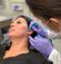A woman with long dark hair reclines in a medical chair as a professional wearing gloves draws lines on her face with a green marker, possibly preparing for a cosmetic procedure. The professional is focused on the woman's face, which has marker guidelines.