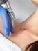 A close-up of a person's collarbone area with a small heart tattoo is being treated with a tattoo removal device. The person operating the device is wearing a blue glove. The area around the tattoo is slightly reddened. A white towel is visible in the background.