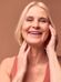 A smiling older woman with blonde hair touches her face with both hands. She is wearing a peach-colored sleeveless top and her expression is joyful. The background is a solid, warm brown color.