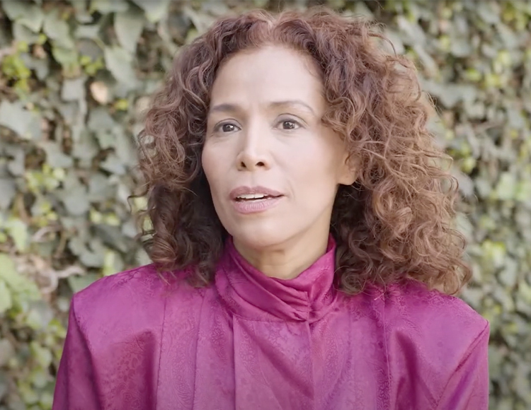 A person with curly brown hair is pictured outdoors in front of a leafy green background. They are wearing a bright magenta top and looking slightly off to the side with a neutral expression.