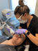 A woman lies on a treatment table with protective goggles on while a clinician uses a Halo laser device on her face. The clinician wears a mask, gloves, and protective eyewear, focusing on the woman's facial treatment in a medical or aesthetic clinic setting.