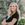 A woman with long, wavy blonde hair stands outside with arms crossed. She is wearing a black top and smiling. The background features greenery with blurred leaves.
